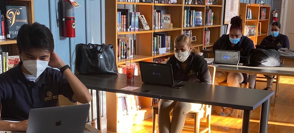 四名学生，一男三女，在图书馆学习。每个人都在自己的办公桌旁，相距6英尺，戴着面具，在笔记本电脑上工作。
